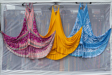 Three colorful female dresses hanging on the outside of a farmer's market booth. The cotton dresses...