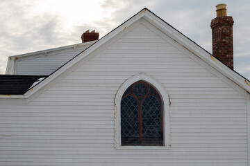 The exterior roof peak at a white wooden church with a dark colored glass gothic style window. The clerestory window has an oval top. There's a chimney on the roof of the traditional building.