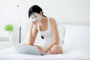Beautiful young asian woman with sheet facial mask sitting working on laptop computer on bed at bedroom, beauty girl applying face mask for skincare of wrinkle at home, skin care, health and wellness.