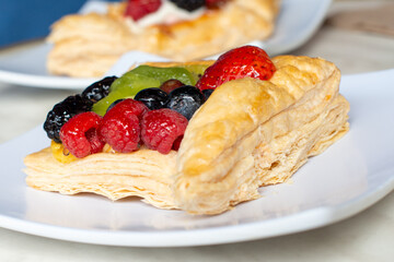 A square fruit filled French puff pastry on a white square plate. The cream and custard filled dessert have strawberries, kiwi, and blackberries on top with a sugar glaze. The pastry is flaky.	