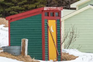 A small yellow and green building with a wooden door. The outhouse or outdoor toilet is in a meadow with white snow. There's a half moon painted on the door. The eco friendly toilet has a door latch. - Powered by Adobe