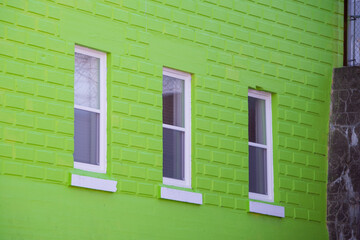 A vibrant lime green block exterior wall of a building with three long casement windows. There's white trim around the closed glass windows. The window sill is white metal.