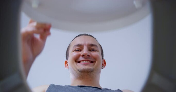 Young man lifts the lid of the toilet, looks in there, says something smiling and closes it back, the view from the inside from bottom up. Guy saw something interesting in toilet bowl