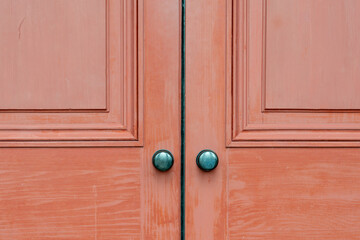Two red wooden doors to a house with two vintage round metal doorknobs. The exterior panel doors show wood grain and worn patterns around the handles. The retro style of doors is hardwood.