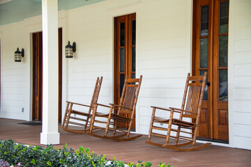 Three vintage brown wooden rocking chairs are on the veranda of an old white colored country house...