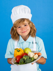 Chef kid boy hold plate with vegetables making fresh vegetables for healthy eat. Portrait of little child in form of cook isolated on blue background. Kid chef. Cooking process.