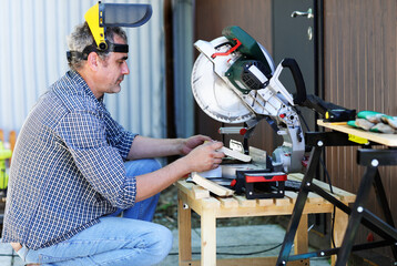 a handyman in a protective helmet works with a miter saw - obrazy, fototapety, plakaty