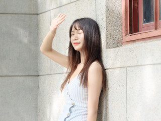 Beautiful Chinese girl with black long hair in sundress leaning against wall raising hand to shade from sun light with eyes closed, daytime relaxation and rest in summer.