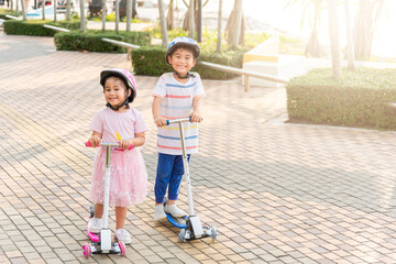 Child riding scooter. Happy Asian little kid boy and girl wear safe helmet play kick board on road in park outdoors on summer day, Active children games outside, Kids sport healthy lifestyle concept - obrazy, fototapety, plakaty