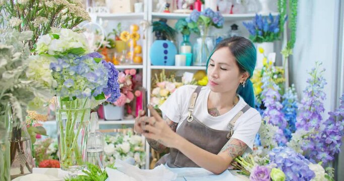 Asian female florist shop owner in apron working in store. Tattooed woman hipster seller taking picture for customer and sending photo to client. E-commerce, SME entrepreneur, Small business online.
