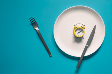 Alarm clock on a white plate with a knife and fork on blue background.