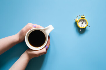 A cup of coffee in female hands and an alarm clock on a blue background.