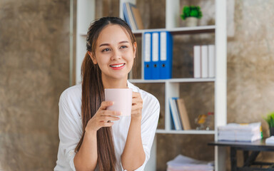 Attractive Asian woman working with laptop, holding coffee cup at home office.