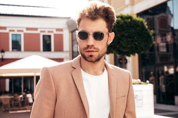 Handsome confident stylish hipster lambersexual model.Sexy modern man dressed in elegant beige suit. Fashion male posing in the street background in Europe city at sunset. In sunglasses