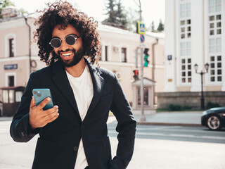 Handsome smiling hipster  model.Sexy unshaven Arabian man dressed in summer suit jacket clothes.Fashion male with long curly hairstyle in the street, using smartphone apps, looking at cellphone screen