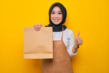 Smiling young Asian Muslim woman barista in white t-shirt apron work in coffee shop hold blank paper takeaway bag mock up and showing thumb up gesture isolated on yellow background. Business startup