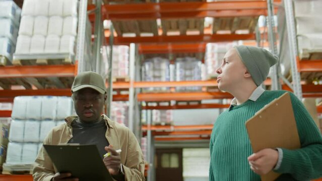 Waist up shot of Caucasian woman and African American man walking in warehouse, talking and taking notes on clipboard while doing inventory together during workday
