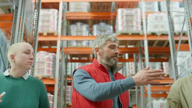 Middle aged manager using digital tablet, pointing on shelves and giving instructions to African American and Caucasian coworkers while walking together through warehouse
