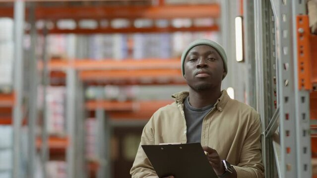 Portrait of young African American man holding clipboard and posing for camera at work in warehouse