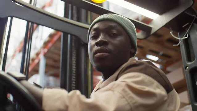Portrait of young African American forklift operator looking on shelves and then posing for camera while working in warehouse