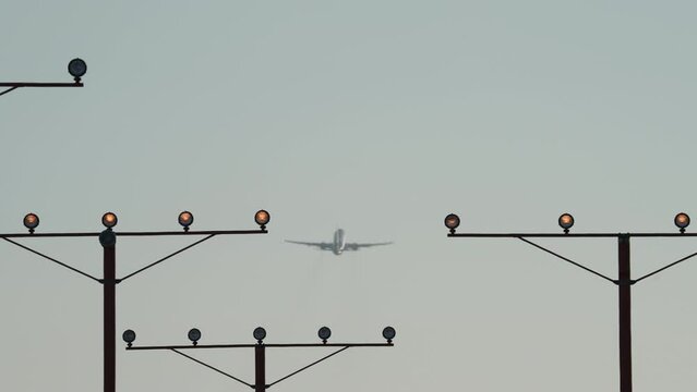 An airliner climbs out after takeoff in the distance, framed by runway approach lights.