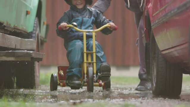 SLOW MOTION, FUNNY shot of a child riding a colourful trike through a muddy puddle