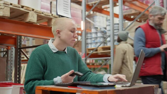 Arc shot of female manager scanning barcodes and using on laptop while working at desk in warehouse
