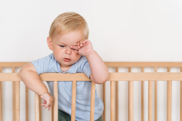 Portrait of beautiful cute little 11 months old sleepy tired baby boy in blue clothes rubbing eyes falling asleep standing in bed leaning on bumpers isolated on white background. Time to nap