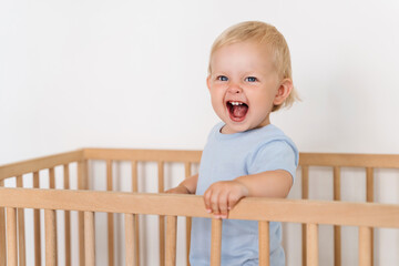 Ten month old baby standing in crib with cheerful excited facial expression, having fun trying to...