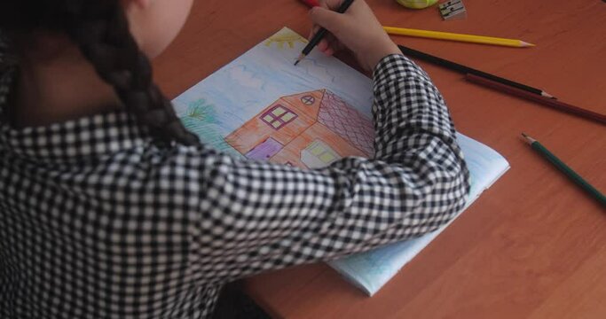 Schoolgirl in a plaid dress with brown hair braided in pigtails sitting at a table in a classroom and drawing a picture of house in an album using color pencils.  