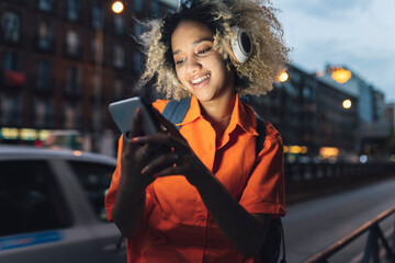 Smiling woman with headphones using smart phone on street at night
