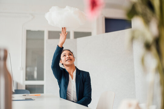 Happy businesswoman sitting at desk touching cloud in office