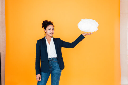 Smiling young businesswoman with levitating cloud in front of yellow wall