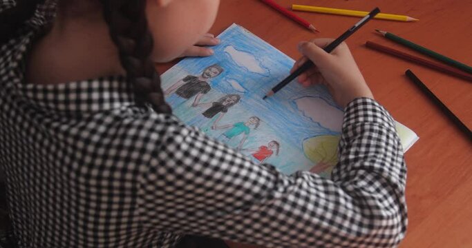 Schoolgirl in a plaid dress with brown hair braided in pigtails sitting at a table in a classroom and drawing a picture of family in an album using color pencils.  
