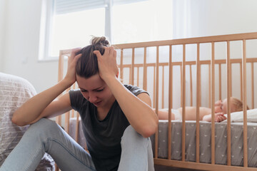 Young mother holding head in hands with desperation, feeling tired and exhausted, crying sitting next to baby sleeping in bed, suffering postnatal depression in need of psychological treatment - obrazy, fototapety, plakaty