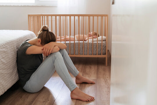 Young mother suffering postnatal depression sitting on floor next to baby napping in bed, resting head on knees, crying, feeling desperate in need of professional psychological support