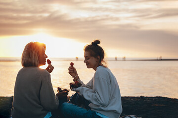 Smiling mother and daughter eating strawberries at beach on sunset