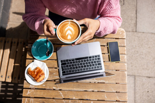 Woman holding coffee cup sitting with laptop at sidewalk cafe on sunny day