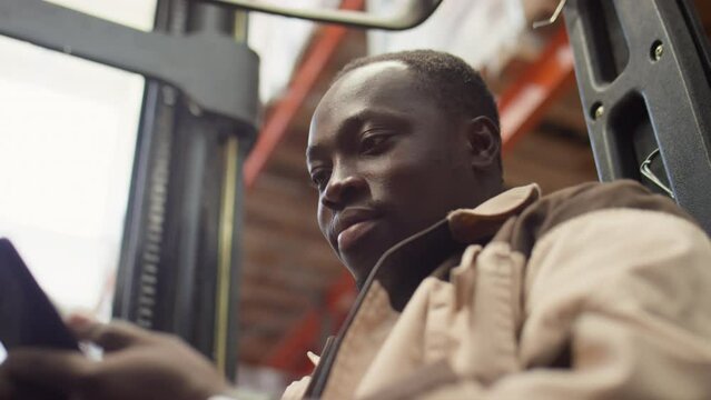 Low angle shot of African American forklift operator typing message on smartphone while working in warehouse