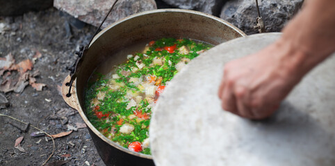 Freshly cooked hot soup in old cauldron on camp fire and man hand with pot cover