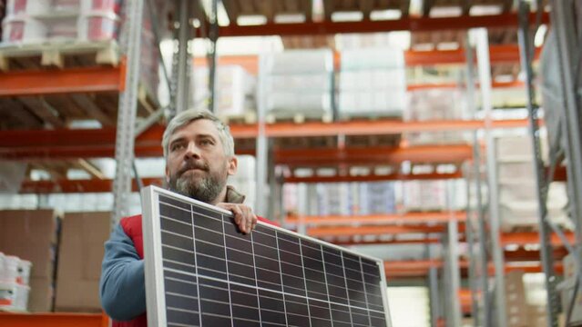 Middle aged worker with gray hair and beard walking along aisle in warehouse and carrying solar panel