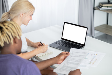 Virtual meeting. Working couple. Digital mockup. Smiling woman making notes while black man checking documents sitting desk with laptop blank screen in light room interior.