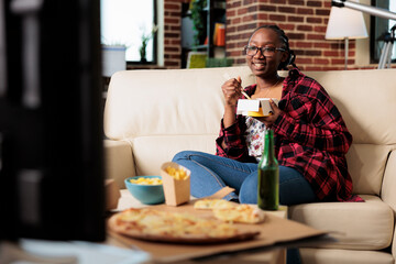Fototapeta na wymiar African american woman eating noodles from takeout delivery package and relaxing on couch with movie on tv. Using choppsticks to eat takeaway fast food meal and drinking beer from bottle.