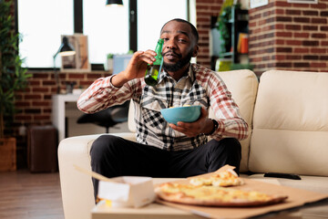 Happy guy drinking alcoholic beer from bottle and holding bowl of chips, watching film on television program. Enjoying beverage with takeaway delivery food and snacks, entertainment.