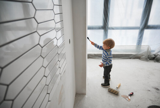 Side view of adorable child painting wall in room under renovation. Cute kid in striped sweater using paint brush while renovating apartment. Concept of home renovation, restoration and childhood.