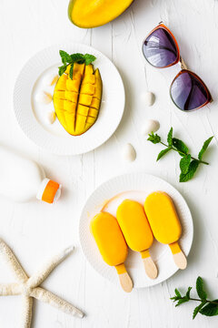 Mango Stileis auf einem Teller und eine frische Mango, eine Sonnenbrille und ein Seestern auf einem weißen Tisch. Flat lay, Sommer.