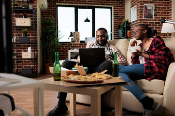 Cheerful man and woman using laptop and eating takeaway food, enjoying meal delivery package to watch movie on television. Watching film and having dinner with fast food and beer.