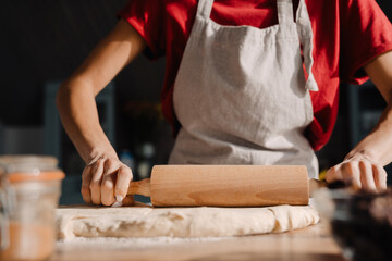 Young woman wearing apron cooking and rolling dough with pin at home
