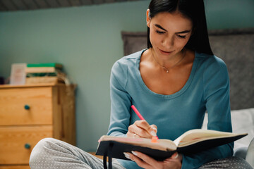 Young hispanic woman writing down notes at organizer in bed at home