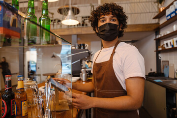 Fototapeta na wymiar Young indian curly barista in face mask working with tablet